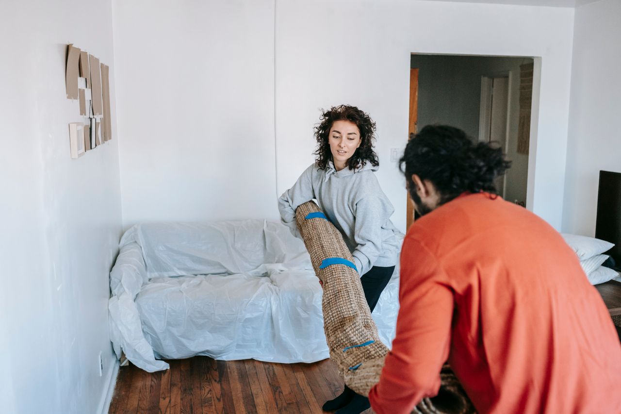 A couple carrying a rolled-up carpet in a sparsely furnished apartment living room.